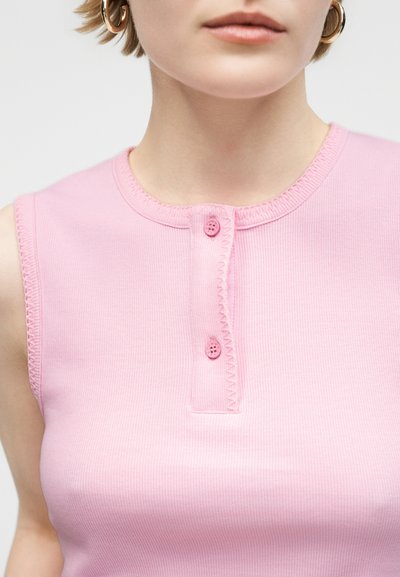 Κοντινή λήψη ενός ατόμου που φοράει ρόζ ριμπ μπλουζάκι χωρίς μανίκια με δύο κουμπιά κοντά στον λαιμό και κρίκους σκουλαρίκια.