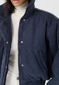 Donkerblauwe jas met ritssluiting en drukknoopsluiting, met een hoge kraag, zijzakken en een zachte textuur. Gelaagd over een witte coltrui.