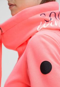 Coral pink hoodie with a high collar, soft texture, featuring black logo patch on the sleeve and cursive text on the collar.