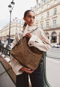 Sac à main en imprimé léopard fabriqué en tissu texturé avec une bordure sombre. Présente une fermeture éclair et une bandoulière. Le modèle le tient dans un cadre urbain.