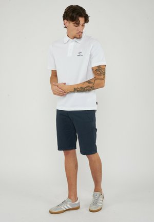 Mann trägt weißes Polohemd, marineblaue Shorts und graue Sneakers, steht mit verschränkten Händen und schaut nach unten auf seine linke Seite.