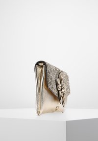 Kulta metallinen laukku, jossa on kimalteleva läppä, teksturoitu pinta, kukkakoristeet ja strassikoristeet. Sivuprofiili valkoista taustaa vasten.