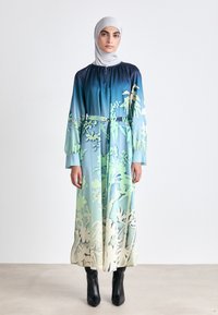 Langes, fließendes Kleid mit einem Ombre-Muster von Dunkelblau zu Hellgrün, das blumige Elemente aufweist. Weicher Stoff und lange Ärmel, in der Taille mit einem Gürtel versehen.
