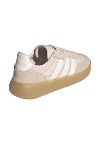 Zapatilla de ante beige con franjas blancas, forma redondeada y suela de goma translúcida. Logo de "Adidas" en el talón en color claro.
