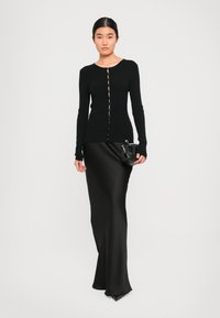 Mulher vestindo um cardigan preto com nervuras e botões, combinado com uma saia longa de cetim preta, segurando uma pequena bolsa preta em frente a um fundo simples.