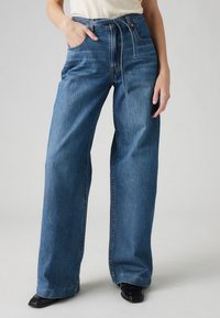 Femme portant un jean bleu taille haute à jambes larges avec une ceinture à cordon, un haut blanc et des chaussures noires à bout carré sur fond uni.