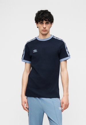 Νεαρός άνδρας με σγουρά σκούρα μαλλιά, που φοράει μπλε σκούρο μπλουζάκι Umbro με φωτεινές γαλάζιες λεπτομέρειες και ταιριαστό γαλάζιο παντελόνι, μπροστά σε μία μονόχρωμη φόντο.