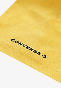 Gul bomuldsstof med ribbet tekstur har sort "CONVERSE" logo og stjernedetalje nær kanten. Nærbillede af designelementerne.