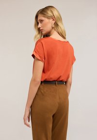 Top ruggine a maniche corte con risvolti, abbinato a pantaloni marroni a vita alta e una cintura scura. Tessuto con texture liscia.