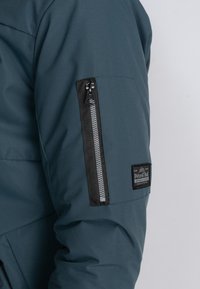 Mørk teal jakke med en glat overflade, der har en lynlås lomme på ærmet med sorte detaljer og et logo-patch.