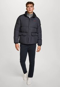 Musta toppatakki, jossa on napillinen edestä, kaksi etutaskua ja pystykaulus, päällä harmaa neulepusero ja tummat housut.