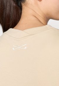 Beige golfsheld met een V-hals en een wit logo van kruisende golfclubs op de bovenrug. Textuurstof met gestructureerde stiksels.