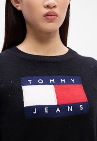 Černý pletený svetr s obdélníkovým logem se sekcemi v červené, bílé a modré barvě a nápisem "TOMMY JEANS" v bílých písmenkách pod ním.