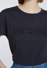 Tmavě námořnické bavlněné tričko s krátkými rukávy a kulatým výstřihem. Obsahuje vyšívaný nápis "EMPOWERED" ve shodné tmavé niti.