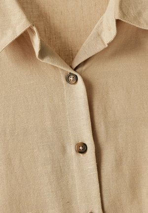 Beige linned skjorte med krave og brune knapper. Stoffet ser tekstureret ud, med synlige fibre og syningsdetaljer.