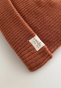 Bonnet en tricot couleur rouille avec une texture côtelée et une petite étiquette crème sur laquelle est brodé "smile LITTLE ONE".