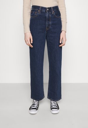 Person iført mørkeblå straight-leg jeans, beige langærmet top og sorte og hvide sneakers, stående på en grå gulv.