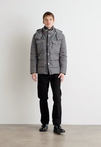Giubbotto imbottito grigio con chiusura a zip e bottoni a pressione, quattro tasche frontali, colletto alto e cappuccio, abbinato a pantaloni e scarpe nere.