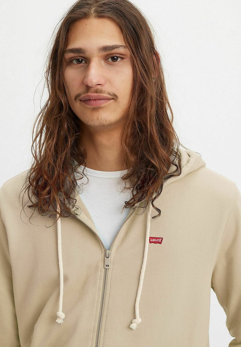Beige Kapuzenpullover mit Reißverschluss und Kordelzügen, aus weichem Material gefertigt. Verfügt über ein kleines rotes Levi's-Logo auf der linken Brustseite. Getragen über einem weißen Shirt.