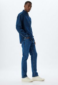 Camisa y pantalones de mezclilla en un tono azul profundo, con detalles de costura. Complementados con zapatillas blancas, de pie contra un fondo neutro.
