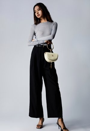 Grauer Langarmoberteil, hoch taillierte schwarze Hose, schwarze Riemchenabsätze und eine runde cremefarbene Handtasche mit strukturiertem Finish und goldenen Akzenten.