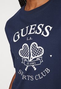 Tummansininen puuvillapaita, jossa on valkoinen graafi, jossa on kaksi sydämenmuotoista tennismailaa ja teksti "GUESS L.A. SPORTS CLUB" alla.