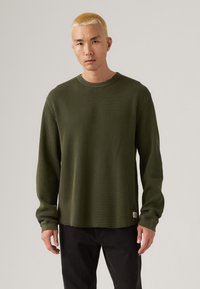 Olijfgroene langemouwen wafelbreiselshirt met een ronde hals en een onbewerkte zoom. Getoond met zwarte broek, heeft een logo patch aan de onderkant.