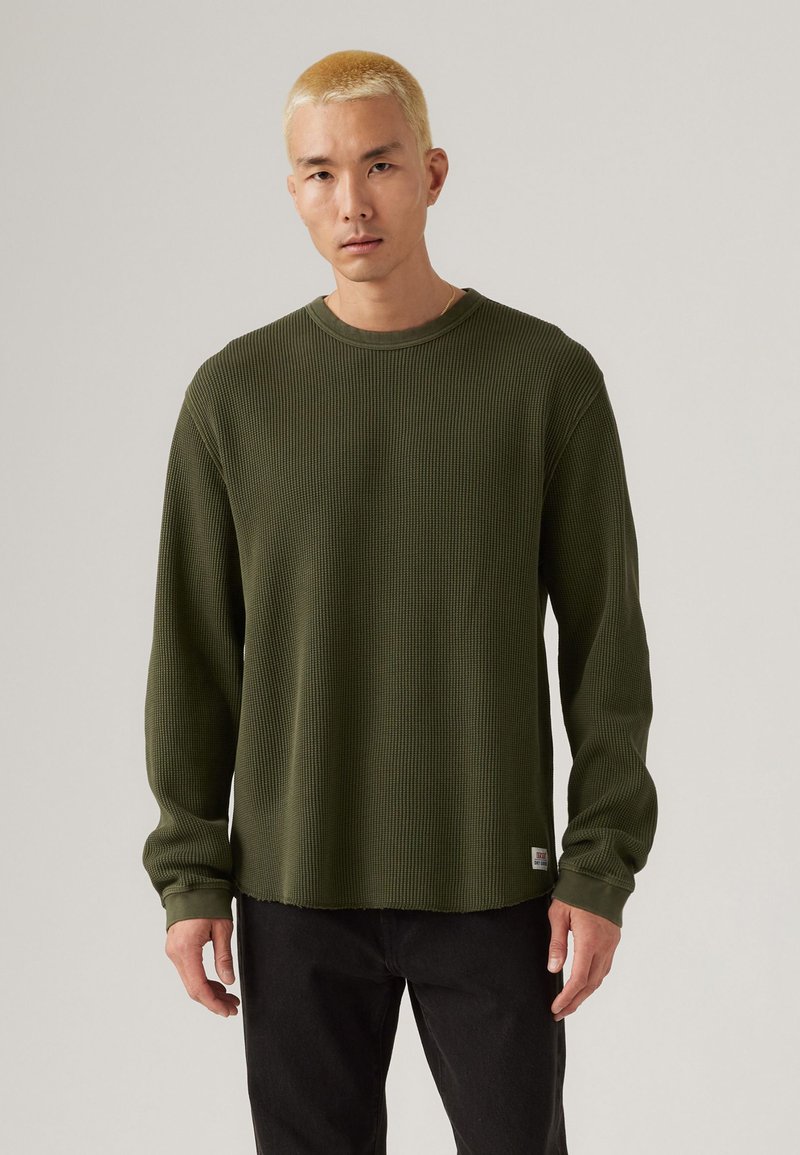 Olijfgroene langemouwen wafelbreiselshirt met een ronde hals en een onbewerkte zoom. Getoond met zwarte broek, heeft een logo patch aan de onderkant.