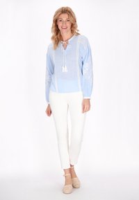 Blusa azzurra chiara con motivi floreali ricamati, maniche arricciate e scollo con laccetto. Abbinata a pantaloni bianchi e calzature neutre.