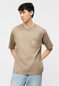 Νέος άνδρας με κοντά μαύρα μαλλιά, που φοράει μπεζ μπλουζάκι με τσέπη στο μπροστινό μέρος και ανοιχτόχρωμα μπλε τζιν, με τα χέρια πίσω από την πλάτη, κοιτώντας προς τα αριστερά.