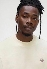 Felpa crema con tessuto testurizzato, scollatura rotonda e logo ricamato color bordeaux sul petto, indossata da una persona con dreadlocks.
