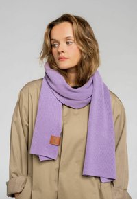 Fioletowy dzianinowy szalik o prążkowanej fakturze, z skórzaną etykietą. Nosi się luźno na beżowej odzieży wierzchniej, podkreślając jego miękkość.