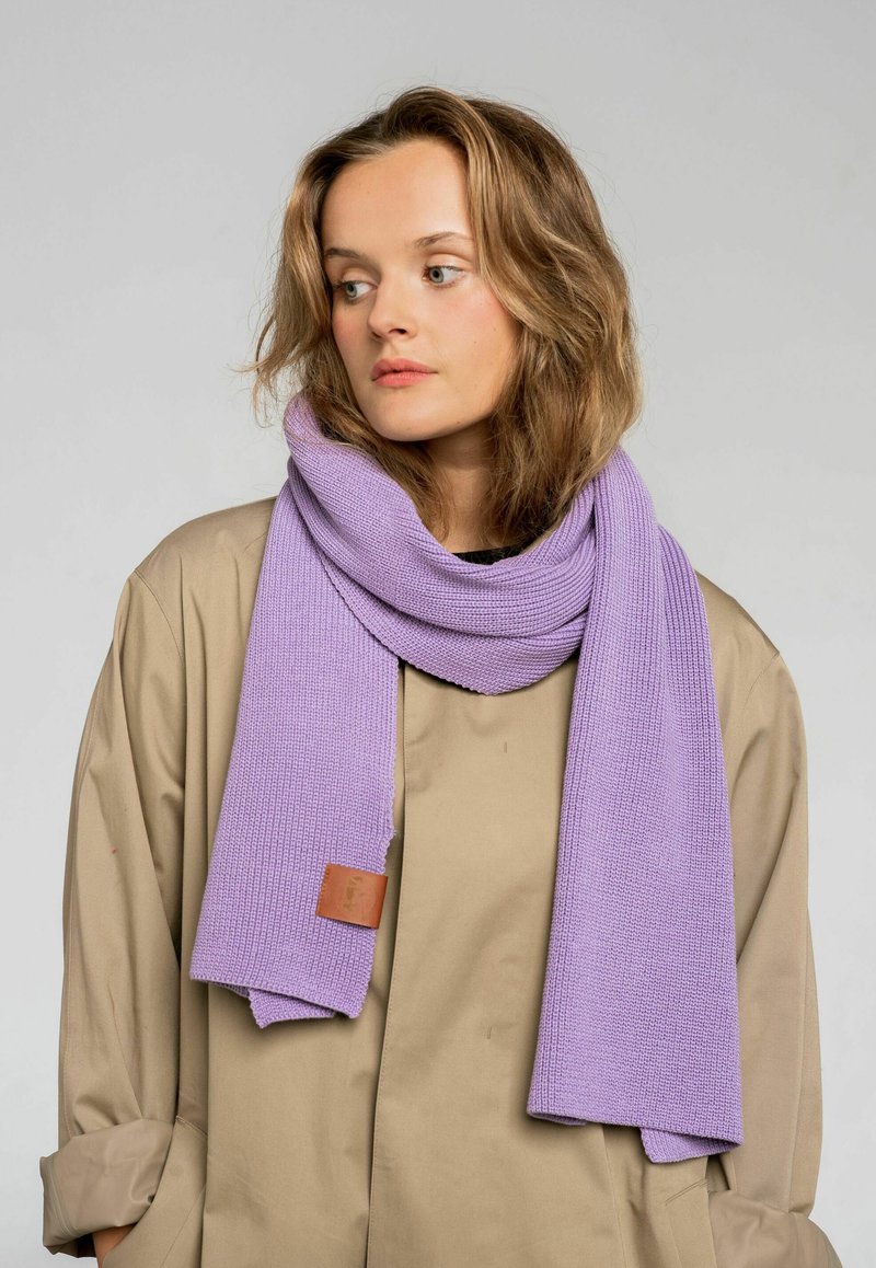 Fioletowy dzianinowy szalik o prążkowanej fakturze, z skórzaną etykietą. Nosi się luźno na beżowej odzieży wierzchniej, podkreślając jego miękkość.