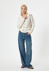Cremfarbener, strukturierter Strickcardigan mit breitem Kragen, Knopfverschluss und kontrastierenden Holzknöpfen, kombiniert mit blauen weit geschnittenen Jeans.