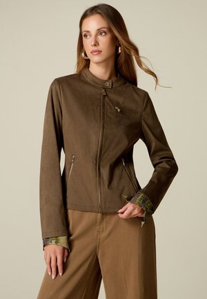 Mujer con cabello largo lleva una chaqueta marrón con cremallera, bolsillos con cremallera y puños de estampado paisley, combinada con pantalones marrones, sobre un fondo liso.