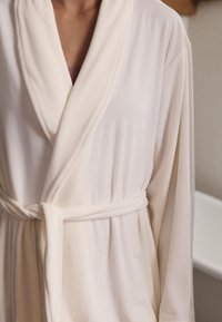 Jasnobeżowy szlafrok z teksturowanymi pionowymi paskami, kołnierzem szalowym i paskiem wiązanym w talii. Wykonany z miękkiego, prążkowanego materiału.