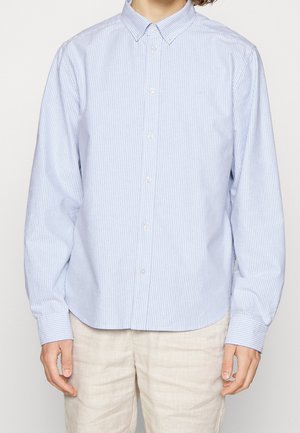 Mężczyzna ubrany w jasnoniebieską i białą koszulę na guziki z pionowymi paskami oraz beżowe spodnie, stojący na tle jednolitego tła.