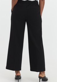 Zwarte wijde broek met een elastische tailleband, voorzien van zijzakken en een soepele textuur. De zoom is iets taps toelopend.