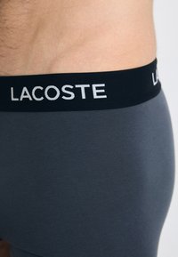 Boxer-briefs gris avec une ceinture élastique noire arborant le logo "LACOSTE" en blanc. Texture lisse, design ajusté.