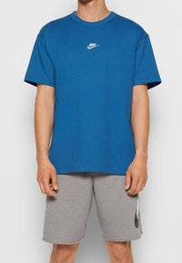 Blå bomullströja med kort ärm, rund hals, och liten vit Nike-logotyp på bröstet, tillsammans med grå bomullsshorts med en svart Nike-logotyp.