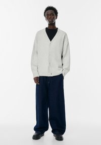 Cardigan lavorato a maglia grigio chiaro con scollatura a V profonda e chiusura con bottoni. Indossato sopra una maglietta nera, abbinato a pantaloni in denim blu scuro larghi. Calzature nere.