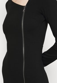 Černé bodycon šaty s dlouhými rukávy, se stříbrným zipem na přední straně a přiléhavou siluetou. Hladká textilie.