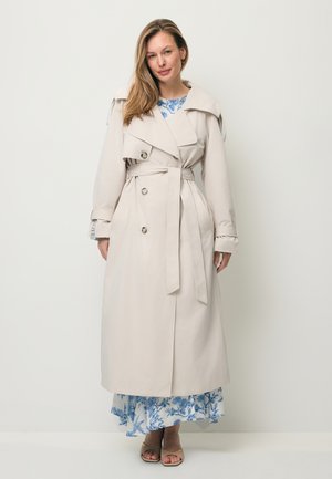 Vrouw draagt een beige trenchcoat met ceintuur over een blauw-witte bloemenjurk en beige sandalen met open tenen, staand tegen een effen witte achtergrond.