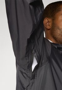 Schwarze, leichte Jacke mit strukturiertem Finish, ausgestattet mit Reißverschluss-Seitenbelüftungen und einem hohen Kragen, die ein elegantes Design und moderne Funktionalität präsentiert.