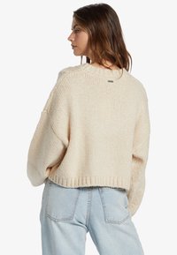 Pull court crème avec un motif tricoté texturé, des manches larges et un ourlet côtelé, comportant une petite étiquette au dos du col.