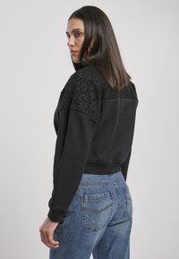 Felpa nera con pannelli in pizzo testurizzato sulle spalle, abbinata a jeans di denim blu chiaro. La vestibilità è rilassata con polsini a coste.