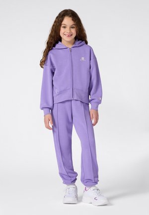 Ragazza sorridente con lunghi capelli ricci, che indossa una felpa a zip color lavanda e pantaloni coordinati, con sneakers bianche, su uno sfondo neutro.