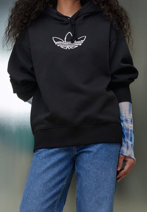 Osoba ubrana w czarną bluzę z kapturem z białym logo Adidas, niebieską, batikową, długą warstwę pod spodem oraz niebieskie dżinsy, stojąca na tle mlecznego szkła.