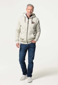 Lichtgrijze rits hoodie met een trekkoord, zakken aan de voorkant en een Union Jack-patch; gecombineerd met blauwe jeans en witte sneakers.