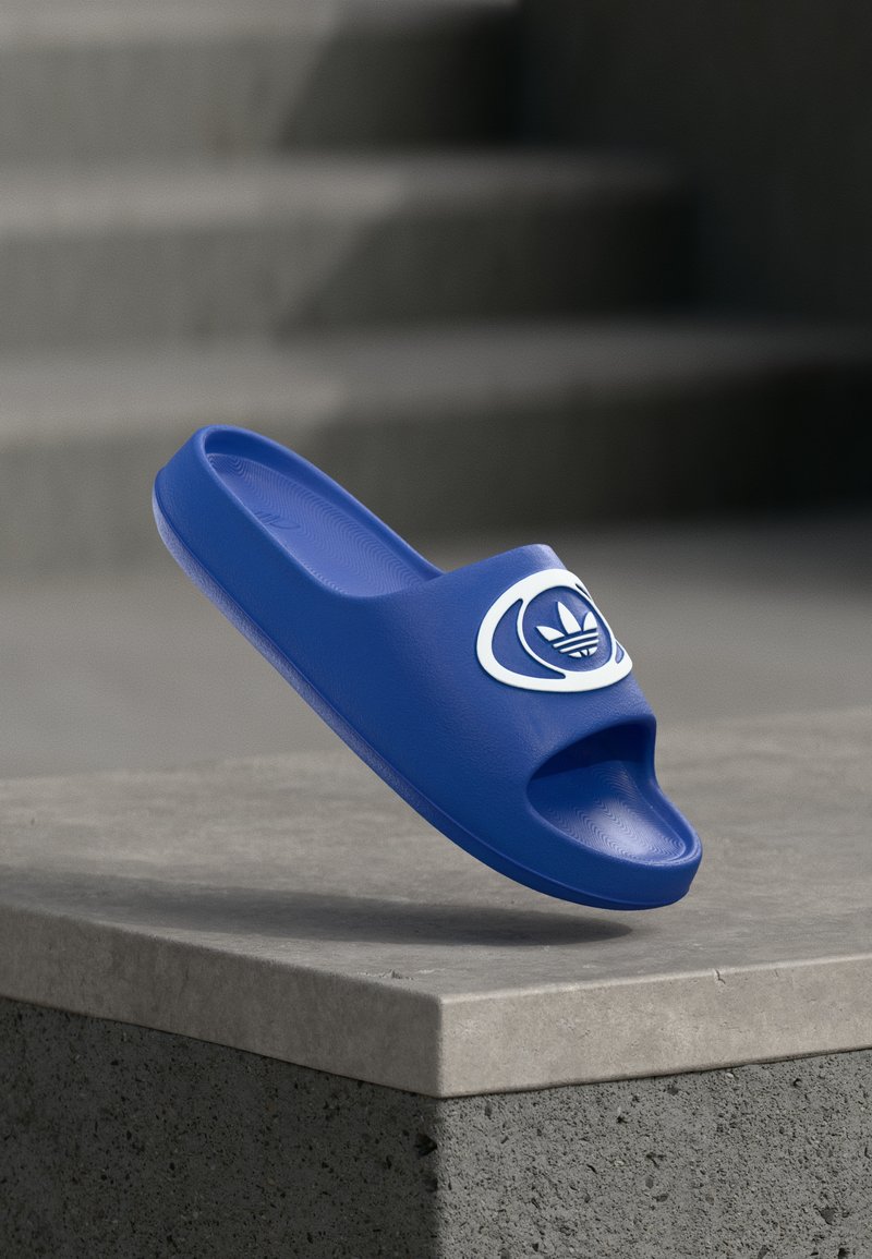 Modrá nazúvacia šľapka vyrobená z hladkého syntetického materiálu, s otvorenou špičkou a bielym logom na remienku.